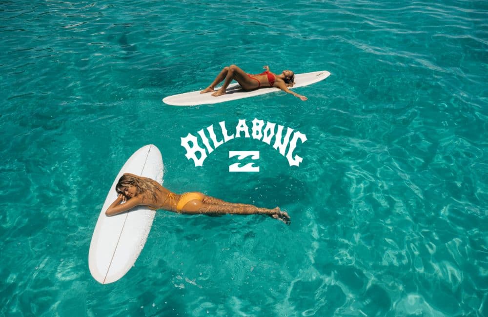 An overhead shot captures two women floating on surfboards in crystal clear, turquoise water.
