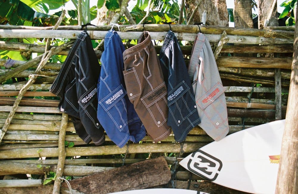 A line of five pairs of boardshorts is hanging on a rustic, woven bamboo or wood fence outdoors.