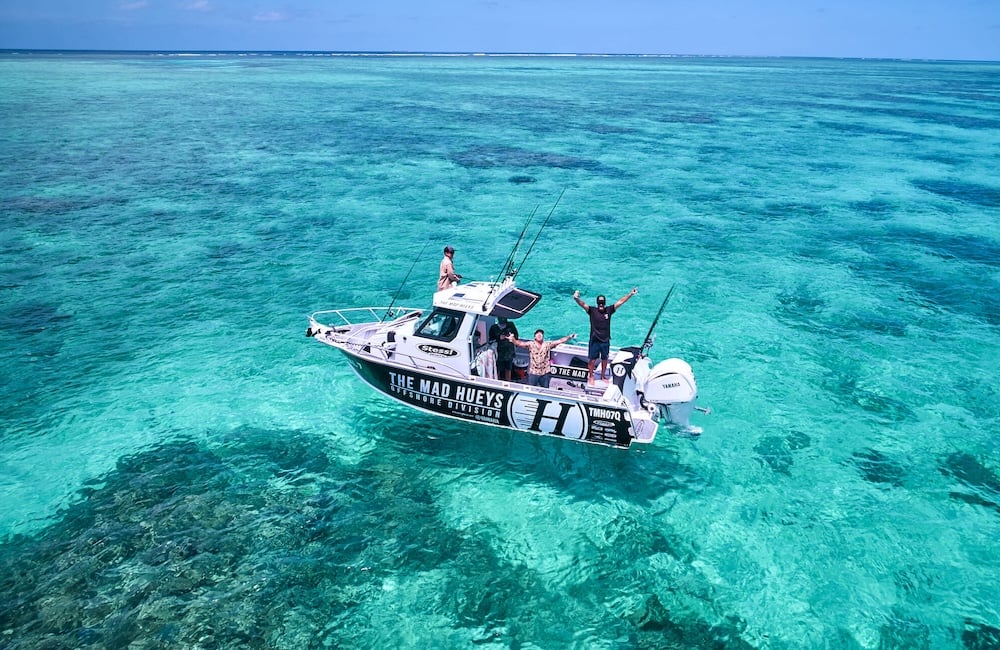 A small, white fishing boat named "THE MAD HUEYS OFFSHORE DIVISION" floating in strikingly clear, shallow turquoise ocean water.